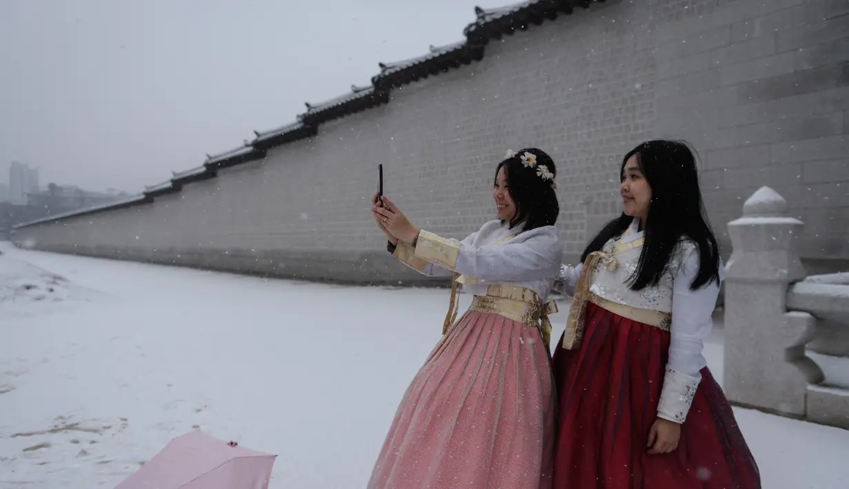 Dua wanita mengenakan gaun hanbok tradisional berselfie di sepanjang dinding istana Gyeongbokgung saat hujan salju di pusat kota Seoul, Korea Selatan (13/12). (AFP Photo/Ed Jones)