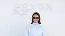 Camila Mendes mengenakan kemeja biru muda dengan tali leher pita, dipadukan rok lipit abu-abu berbelah, tas Prada marun, dan heels senada [dok. Prada]