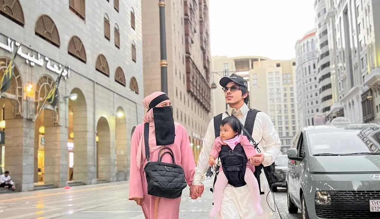 Kembali tampil kompak, kali ini outfit ketiganya tampak lebih cerah. Atta Halilintar menggendong Ameena di depan mengenakan outfit putih, sedang Ameena sendiri tampil kompak dengan Aurelie yang mengenakan outfit berwarna merah muda, kembali dengan burqa berwarna hitam yang menutupi seluruh wajahnya. Foto: Instagram.
