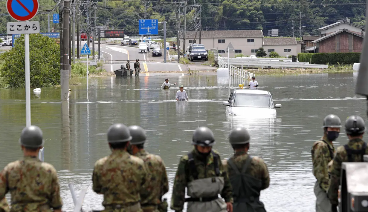 Orang-orang mengarungi jalan yang banjir setelah hujan deras, disaksikan oleh personel Pasukan Bela Diri Darat Jepang, latar depan, di Takeo, Prefektur Saga, Jepang barat daya, Minggu (15/8/2021). (Kyodo News via AP)