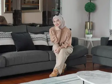 Irish tampil anggun dan tetap kasual dengan sweater cokelat dan hijab yang selaras. Ia duduk manis di sofa, tersenyum sambil menatap ke arah kamera. (Liputan6.com/IG/@ _irishbella_)