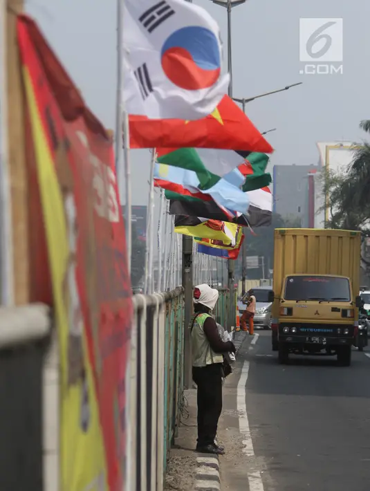 Pedagang berdiri dekat bendera negara peserta Asian Games 2018 di pagar pembatas Jalan Pluit Selatan Raya, Jakarta, Kamis (19/7). Pemasangan bendera bertiangkan bambu itu dianggap justru dapat mempermalukan Indonesia. (Liputan6.com/Arya Manggala)