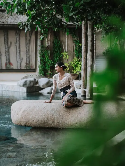 Sebagai aksesori, Mikha mengenakan anting dan cincin emas yang mempercantik penampilannya. Rambutnya ditata rapi dengan sanggul modern, dihiasi bunga kamboja putih di sisi kanan, menambah aura klasik khas Bali. Mikha melengkapi gaya anggunnya dengan tas anyaman mini berwarna krem dan sepasang selop bordir yang tampak nyaman namun tetap menawan. [@miktambayong].