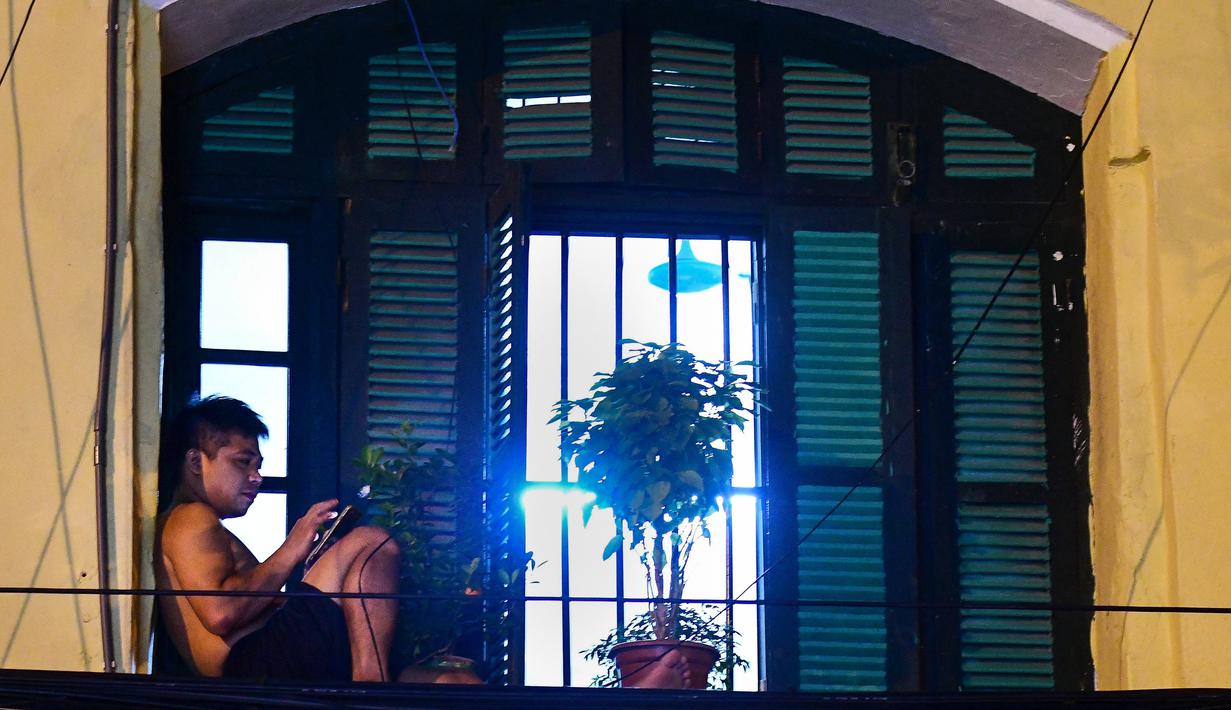 Gambar pada 1 September 2021 menunjukkan seorang pria memeriksa ponselnya sambil duduk di balkon rumahnya di Hanoi, Vietnam, selama lockdown Covid-19. Menghadap ke jalan yang sepi, balkon kecil mereka menjadi saksi bisu terkait aktivitas yang dilakukan oleh masing-masing keluarga. (Nhac NGUYEN/AFP)