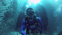 Seorang penyelam merekam pengalamannya menyelam membelah kerumunan ikan. 
