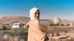 Anggunnya Citra Kirana mengenakan hijab dan dress panjang berwarna putih dipadu dengan syal krem dengan latar belakang balon udara dan padang ilalang di Cappadocia, Turki. (Liputan6.com/IG/@citraciki)