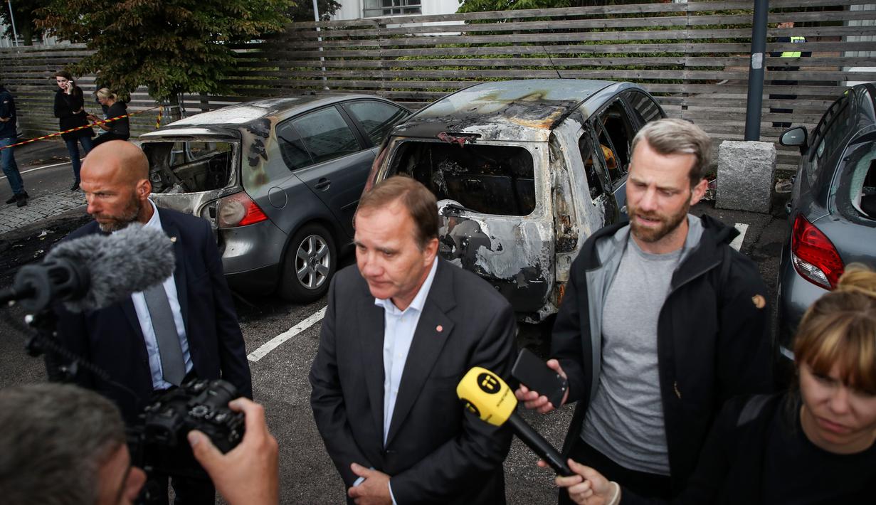 PM Swedia Stefan Lofven memberikan keterangan dengan latar belakang mobil yang hangus dan rusak di kota Gothenburg, Selasa (14/8). Menurut laporan, kelompok gangster menyiram bensin ke atap mobil lalu membakarnya. (Henrik BRUNNSGARD/TT News Agency/AFP)