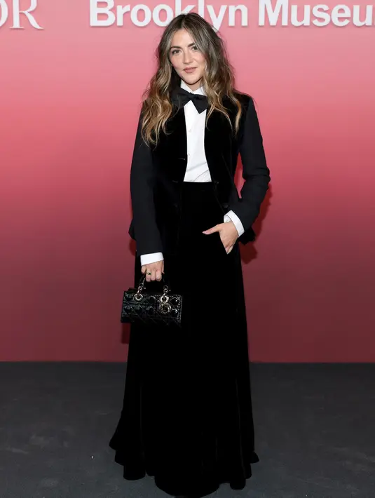 Isabelle Fuhrman mengenakan jaket wol hitam dan beludru Dior Cruise 2024, rok beludru hitam, kemeja katun putih, dan dasi kupu-kupu sutra hitam. Dia juga mengenakan tas dan sepatu Dior. [Dior]