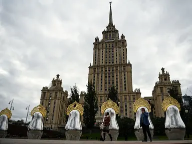 Orang-orang berjalan melewati "benda seni Matryoshki", tujuh boneka bersarang tradisional Rusia "matryoshka" dengan hiasan kepala "kokoshnik", di depan gedung pencakar langit era Soviet "Ukraina" hotel di pusat kota Moskow (9/9/2021).  (AFP/Kirill Kudryavtsev)