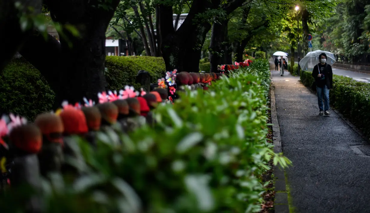 Pejalan kaki dengan mengenakan payung melintasi patung Jizo (kiri) yang terlihat di Kuil Zojoji, Tokyo pada 22 Juni 2020. Jizo adalah pelindung dari anak-anak yang belum sempat lahir atau meninggal dalam kandungan dan saat masih bayi. (Photo by Philip FONG / AFP)