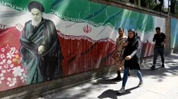 Pejalan kaki melewati lukisan mural yang menggambarkan pendiri Republik Islam Iran Ayatollah Ruhollah Khomeini dan bendera nasional di sepanjang dinding bekas Kedutaan Amerika Serikat (AS) di Ibu Kota Teheran, Iran, Sabtu (22/6/2019). (ATTA KENARE/AFP)