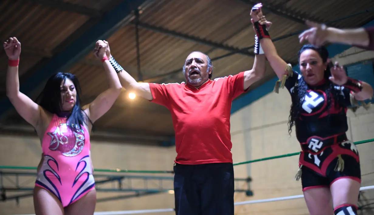 Wasit mengangkat tangan dua pegulat wanita saat bertanding di 2 de Junio Arena di Ciudad Nezahualcoyotl, Negara Bagian Meksiko, Minggu (4/2). Para pegulat saling melepaskan pukulan dan tendangan di atas ring. (Pedro PARDO/AFP)