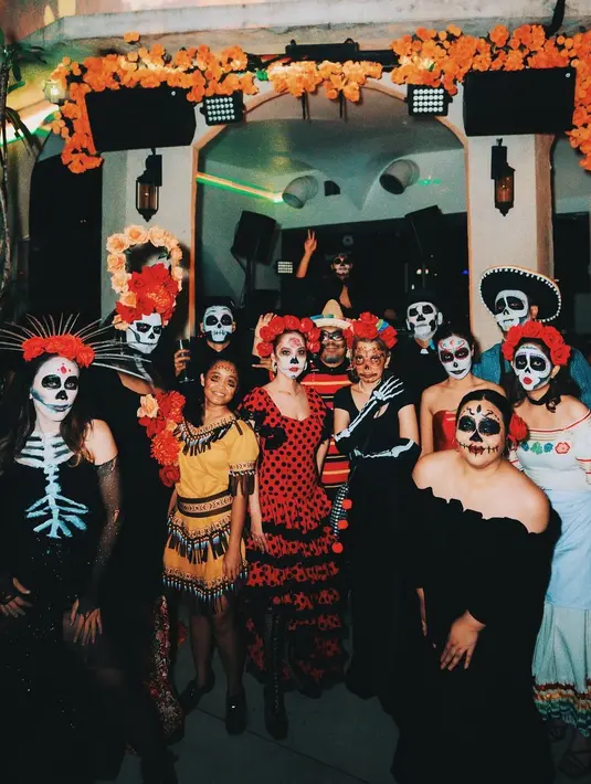 Wajahnya juga dihiasi dengan riasan calavera serupa, dengan sentuhan artistik bunga mawar yang mencerminkan keterkaitan pasangan ini dalam tema mereka. Latarnya yang dipenuhi bunga oranye khas Dia de los Muertos memberikan suasana meriah dan penuh warna, menghidupkan semangat perayaan tradisional tersebut. [@itsmebcl].