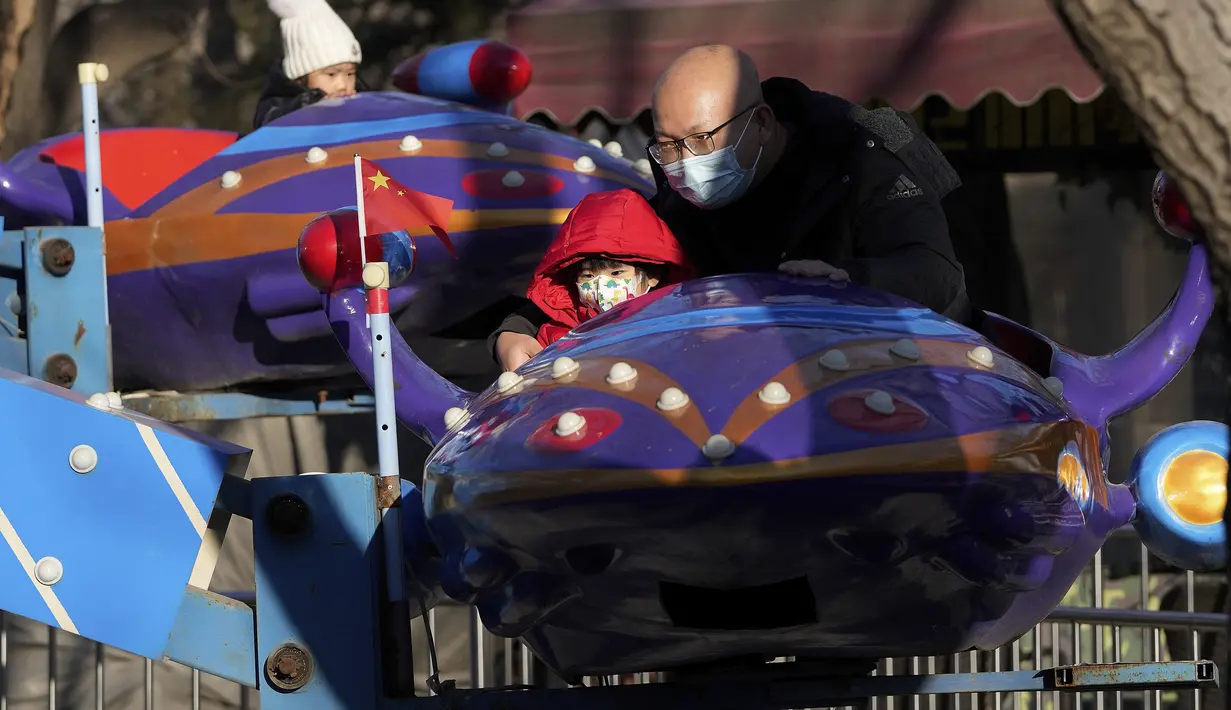 Seorang pria dan seorang anak, keduanya mengenakan masker, mengendarai mesin taman hiburan di Beijing (17/1/2022). Jumlah bayi yang lahir di China terus menyusut tahun lalu, seiring menyusutnya jumlah tenaga kerja. (AP Photo/Andy Wong)