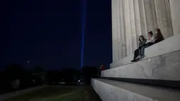 Orang-orang duduk di tangga Lincoln Memorial saat penghormatan 'Menara Cahaya' naik ke angkasa dari dekat Pentagon pada malam peringatan 20 tahun 9/11 di Washington, DC. (10/9/2021). (Anna Moneymaker/Getty Images/AFP)