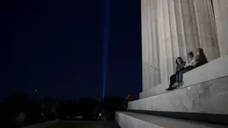 Orang-orang duduk di tangga Lincoln Memorial saat penghormatan 'Menara Cahaya' naik ke angkasa dari dekat Pentagon pada malam peringatan 20 tahun 9/11 di Washington, DC. (10/9/2021). (Anna Moneymaker/Getty Images/AFP)