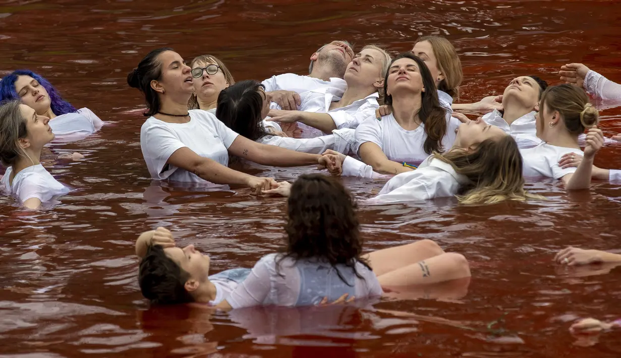 Orang-orang mandi dalam kolam warna merah untuk menandakan darah, selama protes terhadap invasi Rusia ke Ukraina di depan kedutaan besar Rusia di Vilnius, Lithuania, Kamis (7/4/2022). Protes tersebut menyoroti ancaman genosida dari operasi militer Rusia. (AP Photo/Mindaugas Kulbis)