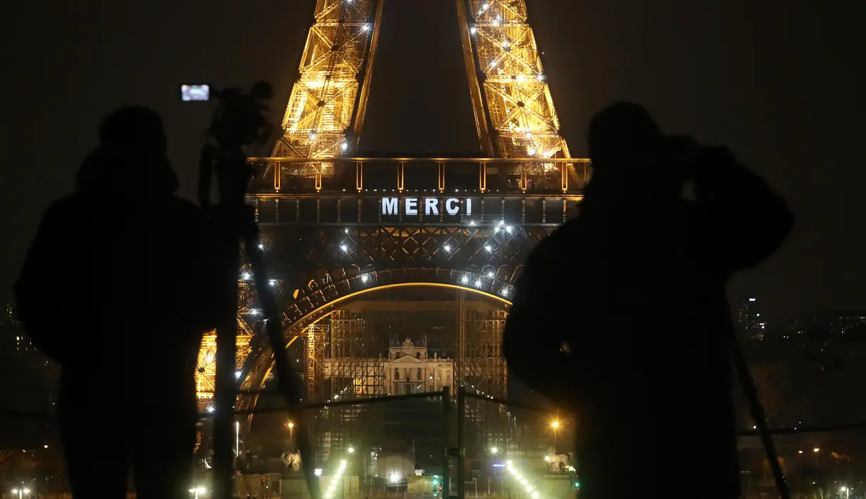 Awak media berdiri di depan Menara Eiffel dengan tulisan 'Merci" (terima kasih) yang terpampang di Paris, Jumat (27/3/2020). Menara Eiffel menampilkan pesan dukungan dan terima kasih kepada tenaga kesehatan di Prancis yang berada di garda depan memerangi pandemi COVID-19.  (AP/Thibault Camus)