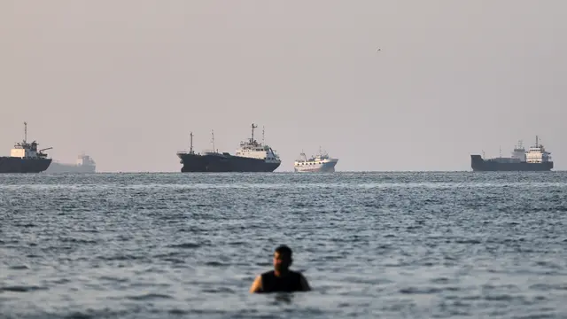 Seseorang berdiri di perairan dangkal saat kapal kargo dan kapal komersial berlabuh di Selat Hormuz di lepas pantai Bandar Abbas, Iran, Senin, 8 Juni 2026. (Amirhosein Khorgooi/ISNA via AP)