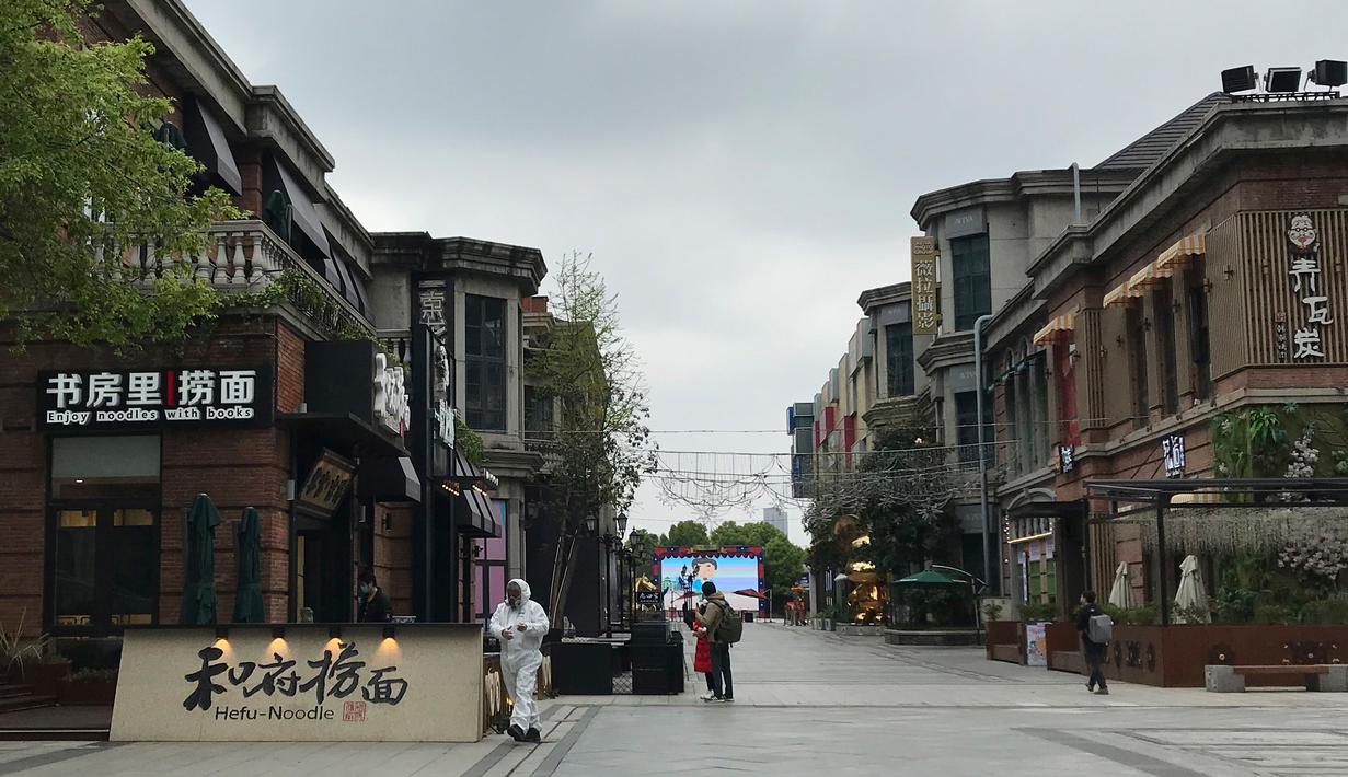Penjaga toko  mengenakan pakaian pelindung menunggu pelanggan di sebuah jalan ritel di Wuhan di provinsi Hubei, China tengah (30/3/2020). Penjaga toko menyiapkan pembersih tangan dan memeriksa tanda-tanda demam pada pelanggan yang datang. (AP Photo/Olivia Zhang)