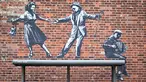 Sebuah karya seni grafiti dari pasangan yang menari dengan pemain akordeon, yang menyandang ciri khas seniman jalanan Banksy, terlihat di dinding di Great Yarmouth di pantai timur Inggris pada 8 Agustus 2021. Karya tersebut belum diklaim oleh Banksy. (JUSTIN TALLIS / AFP)