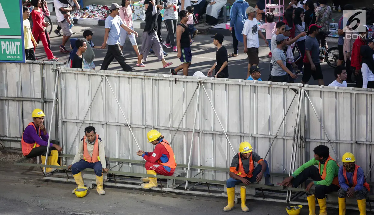 Sejumlah buruh proyek duduk di pintu gerbang dengan latar belakang warga yang sedang beraktifitas saat berlangsungnya Hari Bebas Kendaraan Bermotor (HBKB) di kawasan Bunderan HI, Jakarta, Minggu (24/12). (Liputan6.com/Faizal Fanani)
