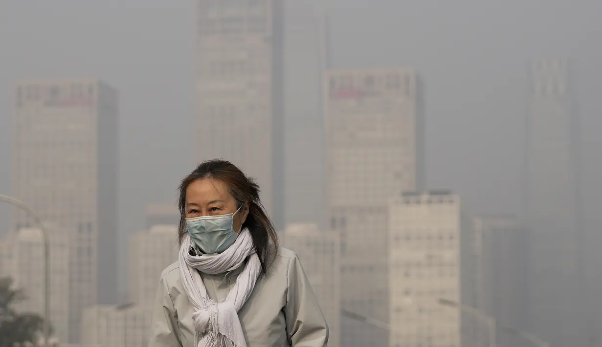 Seorang wanita mengenakan masker untuk melindungi diri dari COVID-19 berjalan di depan gedung perkantoran di Kawasan Pusat Bisnis yang diselimuti kabut polusi di Beijing, Kamis (18/11/2021).  (AP Photo/Andy Wong)