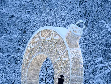 Seorang perempuan berjalan melewati lengkungan ringan yang disiapkan untuk liburan Tahun Baru dan Natal mendatang di depan pohon yang tertutup salju setelah hujan salju lebat di pinggiran Moskow, Rusia pada Selasa (14/12/2021). (Kirill KUDRYAVTSEV / AFP)