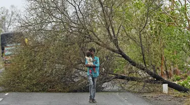 Seorang pria menggendong anak saat berjalan melewati pohon yang tumbang akibat terjangan siklon Tauktae di dekat Amreli, India, Selasa (18/5/2021). Topan Tauktae disebut yang terbesar melanda wilayah itu dalam beberapa dekade terakhir. (Punit PARANJPE / AFP)