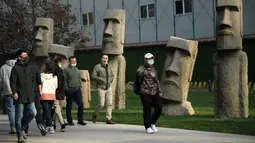 Orang-orang yang memakai masker berjalan melewati replika patung raksasa 'Moai' Pulau Paskah di Beijing pada Rabu (4/11/2020). Pulau Paskah yang berada di Negara Chile terkenal oleh patung kepala yang dipahat dari batu. (Photo by Noel Celis / AFP)