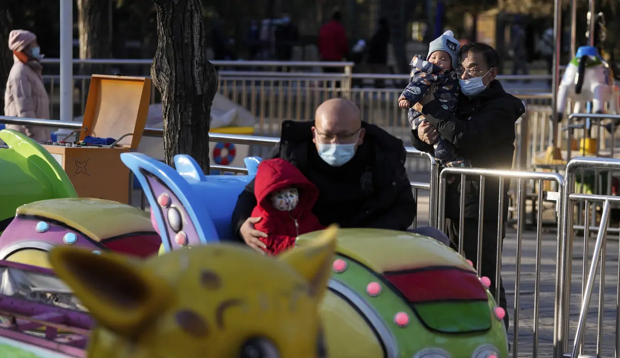 Seorang pria yang mengenakan masker menggendong seorang anak menonton keluarga naik kereta mainan di sebuah taman hiburan di Beijing (17/1/2022). Jumlah bayi yang lahir di China terus menyusut tahun lalu, seiring menyusutnya jumlah tenaga kerja. (AP Photo/Andy Wong)