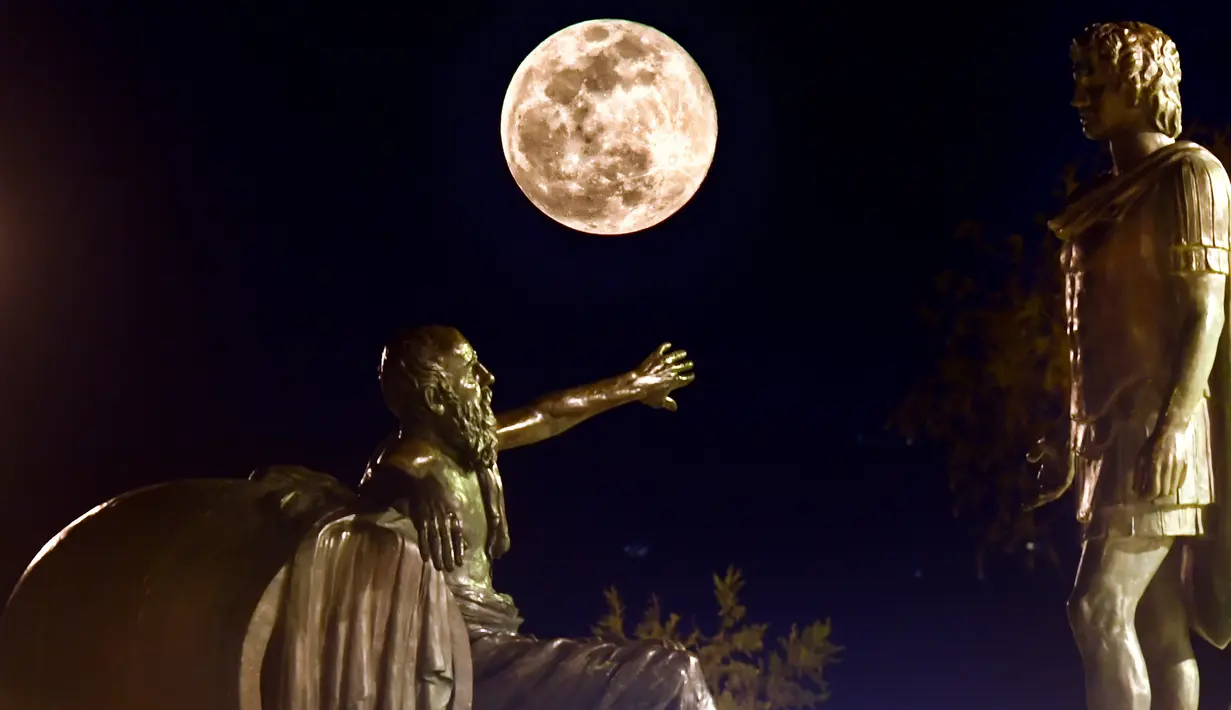 Fenomena super snow moon terlihat di antara patung Alexander Agung (kanan) dan Diogenes of Sinope (kiri) di Corinth, Yunani, Selasa (19/2). Super snow moon menampakkan diri di berbagai belahan dunia. (Valerie GACHE/AFP)