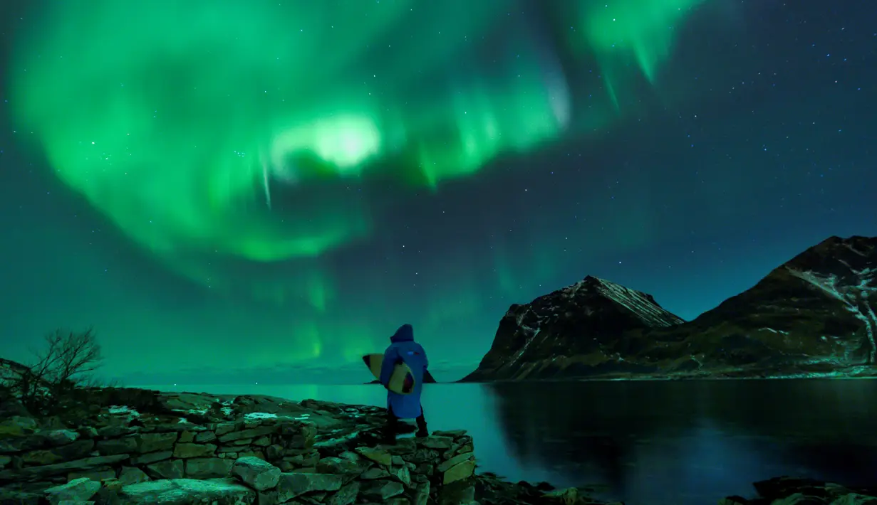 Seorang surfer membawa papannya melihat Cahaya Utara atau aurora borealis di Utakleiv, Norwegia utara (9/3). Biasanya aurora terjadi di daerah di sekitar kutub Utara dan kutub Selatan magnetiknya. (AFP/Olivier Morin)