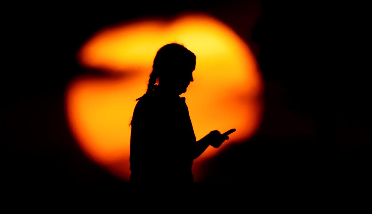 Seorang wanita melihat teleponnya di sebuah taman ketika matahari terbenam di Kansas City, Missouri, Amerika Serikat, Senin (29/6/2020). Matahari terbenam dan matahari terbit lebih cerah dari biasanya belakangan ini karena debu di atmosfer dari awan debu Sahara. (AP Photo/Charlie Riedel)