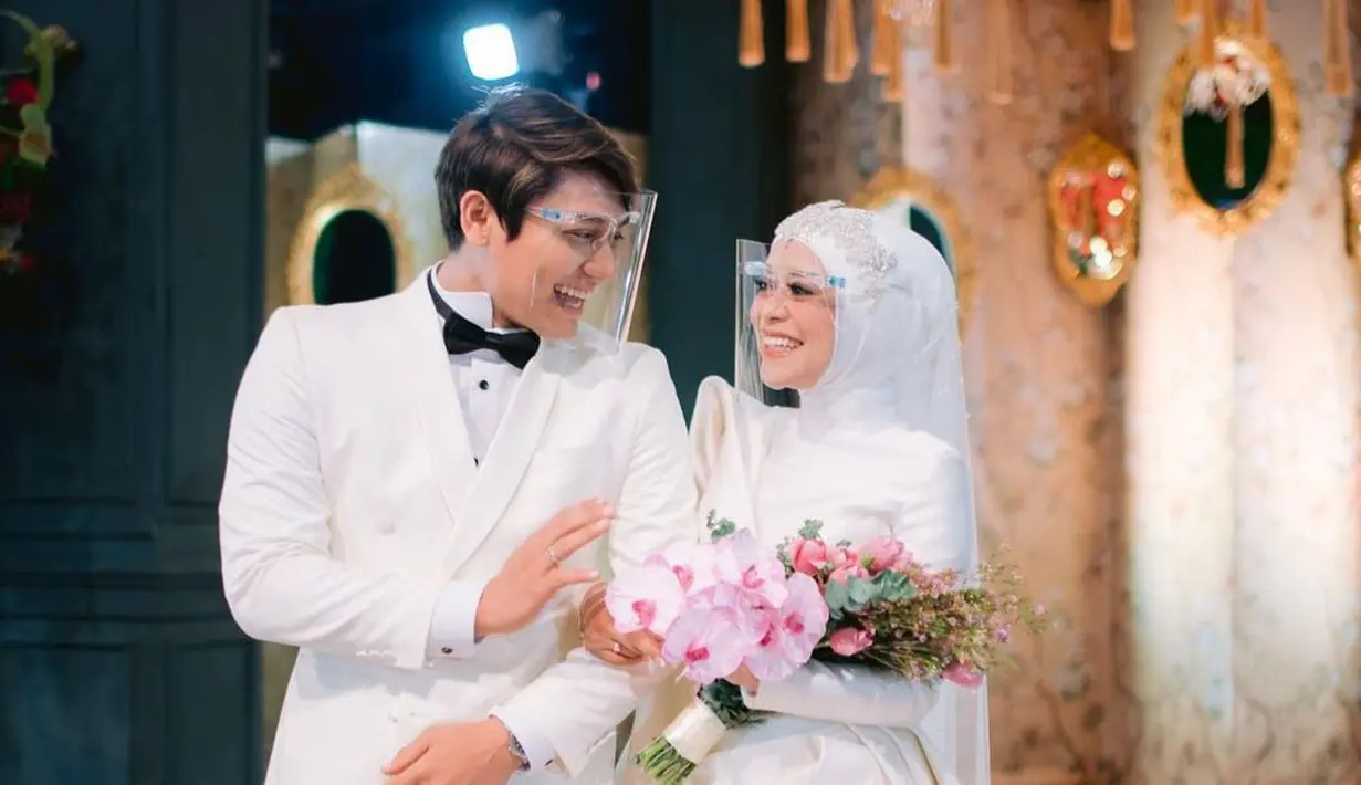 Foto lain menampilkan Rizky Billar mengenakan setelan jas putih dengan dasi kupu-kupu hitam yang dipadu dengan celana panjang hitam. Lesti Kejora pakai gaun pengantin serbaputih berkonsep modern menyesuaikan dengan penampilannya yang berhijab. Pernikahan di tengah pandemi Covid-19 ini menerapkan protokol kesehatan ketat. Keduanya pakai face shield. (Foto: Dok. Instagram @rizkybillar)