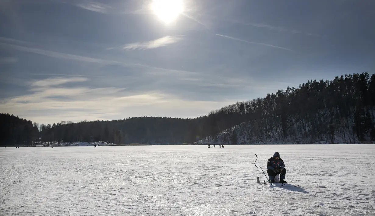 Seorang pria memancing melalui lubang es di danau beku yang tertutup salju dekat Vilnius, Lithuania, Minggu (13/2/2022). Suhu udara 0 derajat Celcius (32 derajat Fahrenheit). (AP Photo/Mindaugas Kulbis)