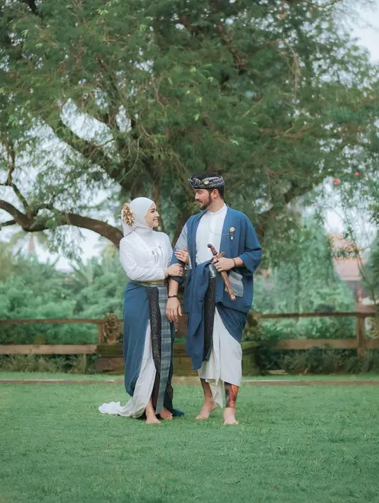 Kompak memakai baju adat nuansa biru, mereka tampak ceria berpose di tengah pemandangan alam. Keduanya tampak bergandengan dan saling menatap, romantis banget.(Liputan6.com/IG/@aryanifitriana24)