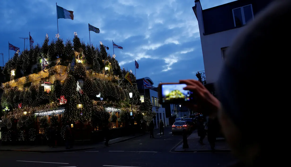 Pejalan kaki mengambil gambar dari bangunan bar The Churchill Arms yang ditutupi pohon natal dan lampu warna-warni di London, Inggris, 20 Desember 2016. Churchill Arms memang dikenal berkat dekorasi Natalnya yang sangat meriah. (REUTERS/Neil Hall)