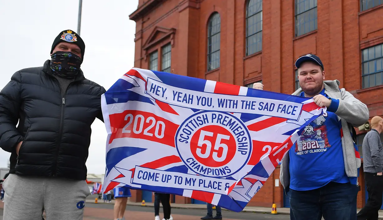 Dua fans Glasgow Rangers membentangkan bendera klub di luar Ibrox Stadium (7/3/2021) untuk merayakan keberhasilan tim kesayangan mereka merebut gelar Liga Utama Skotlandia setelah memimpin 20 poin dari saingan terdekat, Glasgow Celtic. (AFP/Andy Buchanan)