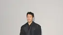 Son Suk-ku tampil dengan trench coat navy panjang yang dililit sabuk kulit lebar di pinggang, dipadukan kemeja dan celana serba hitam [dok. Burberry]