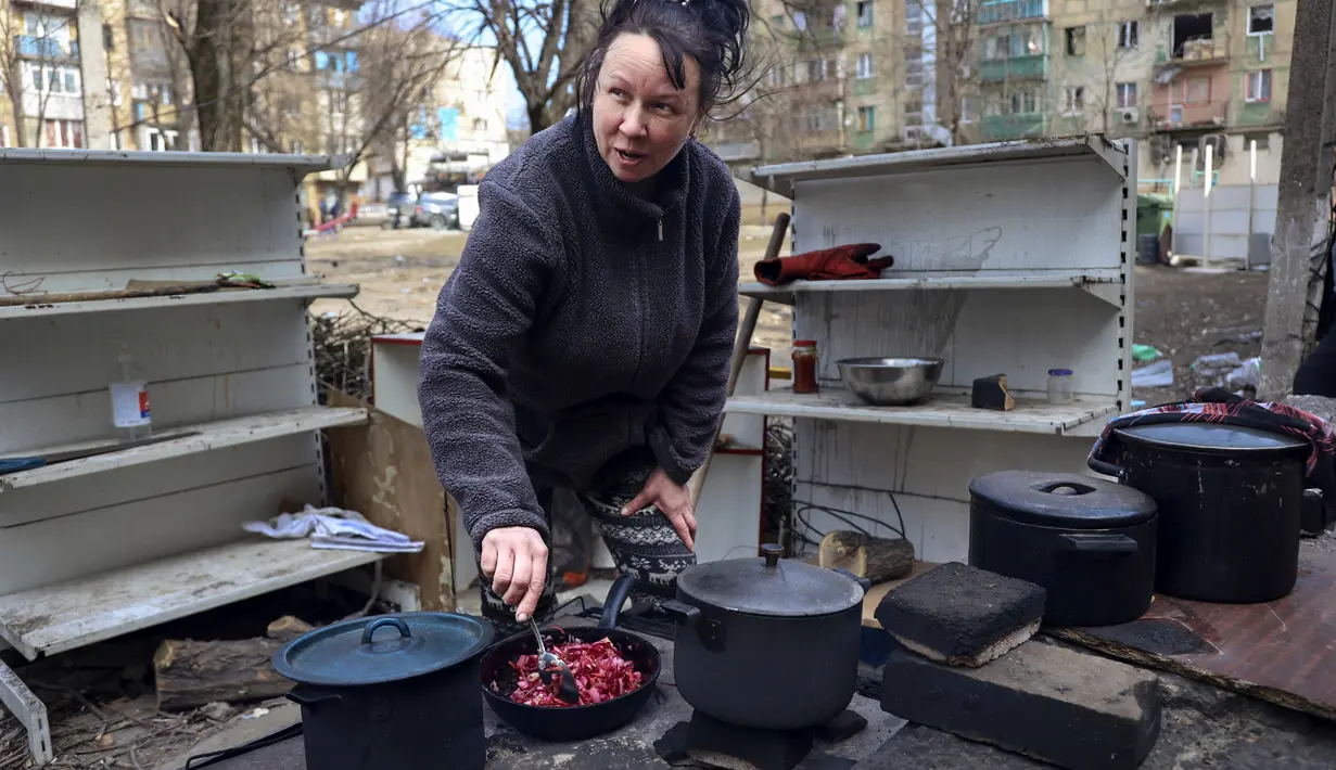Seorang perempuan memasak di halaman gedung apartemen yang rusak akibat tembakan dari pertempuran di pinggiran Mariupol, Ukraina, di wilayah di bawah kendali pemerintah separatis Republik Rakyat Donetsk, pada Selasa, 29 Maret 2022. (AP Photo/Alexei Alexandrov )