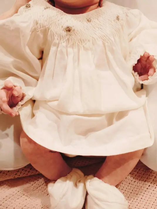 Sementara itu, Baby Soleil tampil menggemaskan dalam gaun putih berpotongan longgar dengan detail kerut dan kaus kaki senada. Busana serba putih tersebut semakin menonjolkan kesan polos dan manis yang identik dengan bayi baru lahir. [@alyssadaguise].
