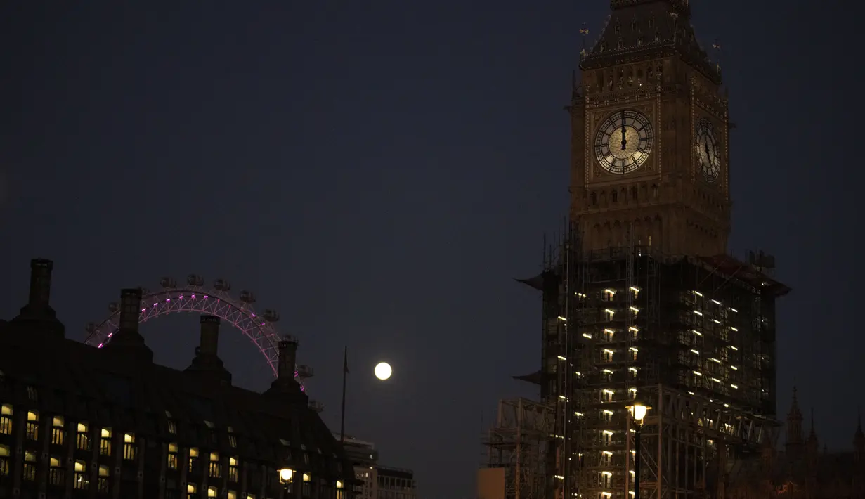 Bulan purnama pertama tahun ini atau Wolf Moon terlihat di belakang London Eye dan tampilan jam saat perbaikan Menara Elizabeth, yang dikenal sebagai Big Ben dan Gedung Parlemen, di London, Senin (17/1/2022). Wolf Moon adalah sebutan untuk bulan purnama pertama di Januari. (AP Photo/Matt Dunham)