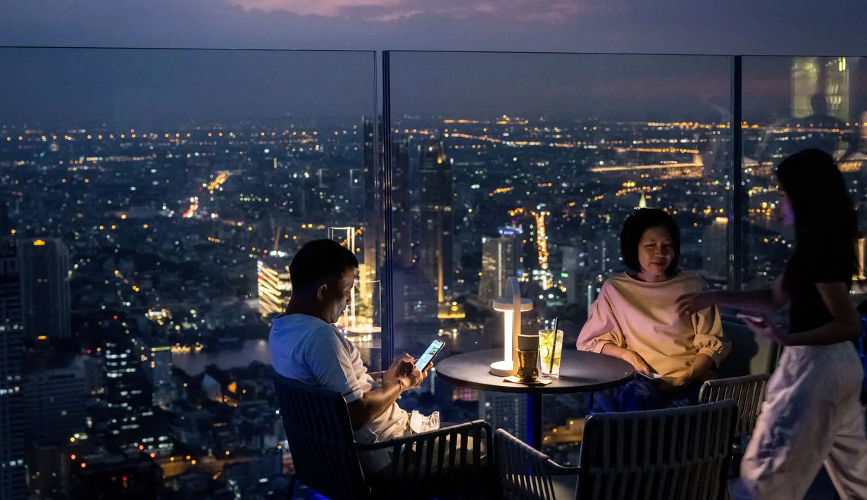 Orang-orang minum dengan latar belakang cakrawala Bangkok di dek observasi gedung pencakar langit King Power Mahanakhon pada 25 Oktober 2021. Gedung tertinggi di Thailand itu juga membuka rooftop bar dan lokasi lantai tembus pandang seluar 63 meter persegi. (Jack TAYLOR / AFP)