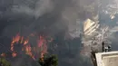 Api membakar pohon di atas bukit saat orang-orang menyaksikan kebakaran hutan di pinggiran kota Voula, di selatan Athena, Yunani, Sabtu, 4 Juni 2022. Kebakaran hutan berkobar di luar kendali di pinggiran Athena, hingga merusak rumah-rumah dan mobil di permukiman warga. (AP Photo/Yorgos Karahalis)