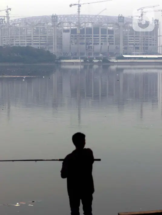 Warga memancing di pinggir Waduk Cincin yang berlatar pembangunan Jakarta International Stadium (JIS), Sabtu (19/6/2021). Pembangunan JIS sudah memasuki tahap pemasangan rangka atap dengan bentang mencapai 269 meter dan ditarget selesai Desember 2021. (Liputan6.com/Helmi Fithriansyah)