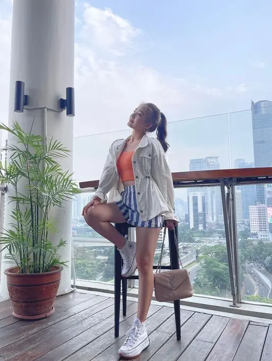 Next, crop top warna oranye dengan rok stripes menjadi pilihan Natasha Wilona. Dia memadukan outfit tersebut dengan parka putih dan sneakers senada. (Instagram/natashawilona12).