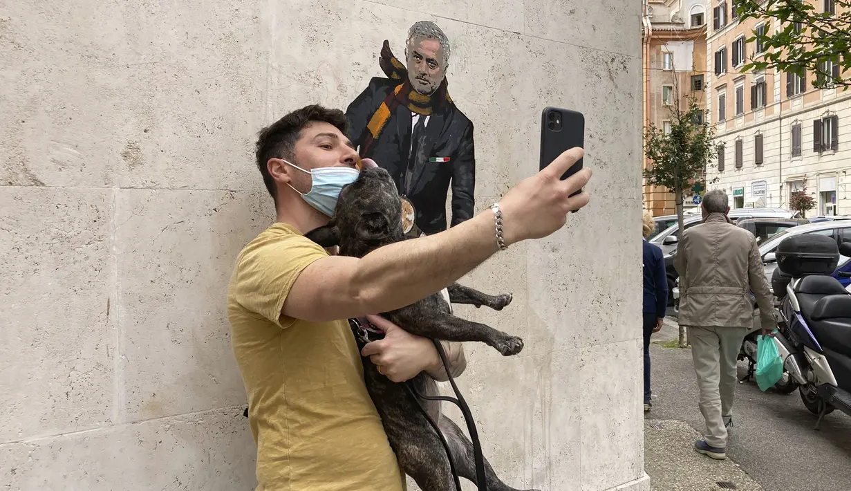 Seorang pria berselfie di dekat mural pelatih Jose Mourinho mengendarai skuter Vespa berhias lambang AS Roma di lingkungan Testaccio,  Roma (7/5/2021). AS Roma mengontrak pelatih Jose Mourinho sampai tahun 2024. (AP Photo/Gregorio Borgia)