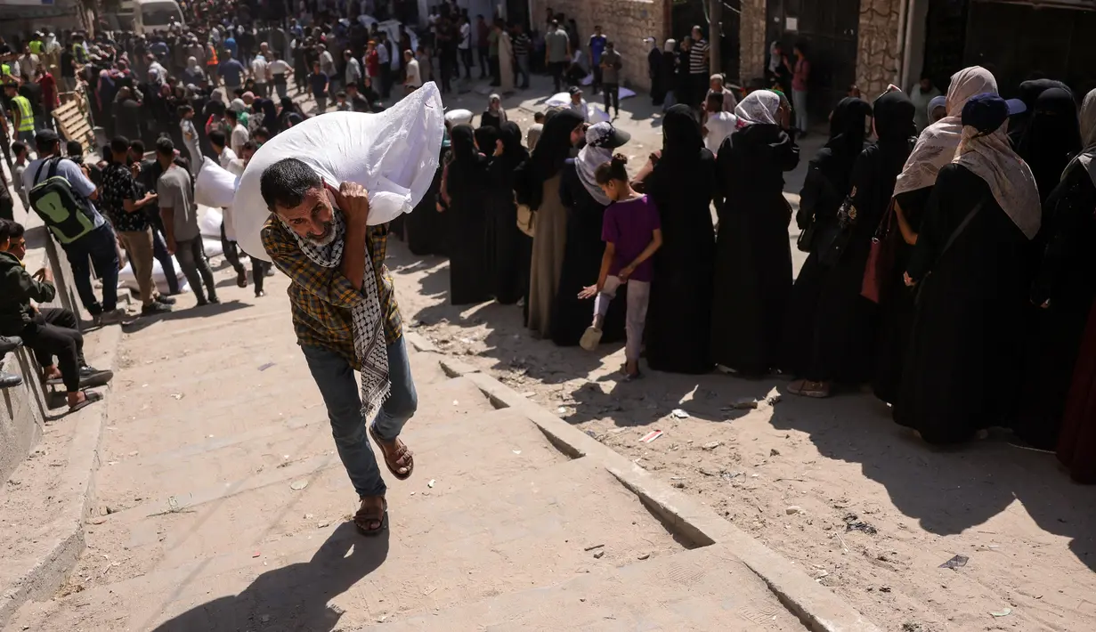 Seorang pria Palestina membawa sekantong tepung, sementara yang lain menunggu di depan titik distribusi yang didirikan oleh Save Youth Future Society (SYFS), yang bekerja sama dengan Program Pangan Dunia PBB (WFP), di Kota Gaza pada 26 Juni 2025. (BASHAR TALEB/AFP)