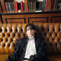 Dalam sebuah momen photoshoot, Jungkook BTS terlihat layaknya bangsawan. Ia mengenakan high-neck innerwear satin berwarna putih, yang memiliki detail kerut dan dasi. Untuk menambah kesan layered pada penampilannya, ia mengenakan long-outerwear yang juga berbahan satin. Ia padukan keseluruhan penampilannya dengan celana panjang hitam dan beberapa aksesori seperti cincin dan anting. Foto: Instagram.